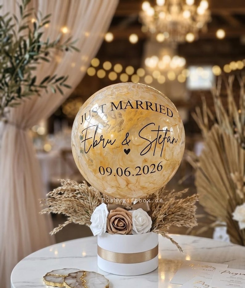 以下が含まれることがあります： 金色の花びらが入った透明なバルーン。黒い文字で「JUST MARRIED」「Ebru & Stefan」「09.06.2026」と書かれています。バルーンは、白と金のバラ、ドライパンパスグラスで飾られた白い円筒形の箱の上に設置されています。