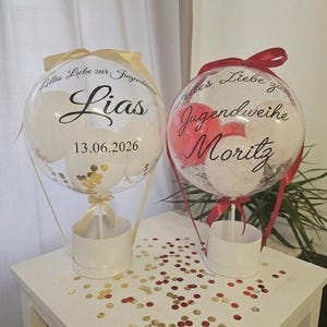 Op de afbeelding: Twee doorzichtige, bolvormige ballonnen gevuld met kleinere witte en rode ballonnen en gouden confetti. De linkerballon bevat de tekst "Alles Liebe zur Jugendweihe", "Lias" en "13.06.2026". De rechterballon bevat "Alles Liebe zur Jugendweihe" en "Moritz".