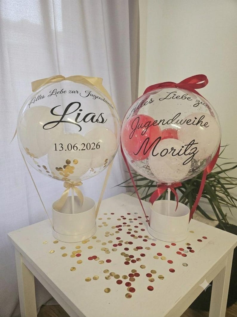 Op de afbeelding: Twee doorzichtige, bolvormige ballonnen gevuld met kleinere witte en rode ballonnen en gouden confetti. De linkerballon bevat de tekst "Alles Liebe zur Jugendweihe", "Lias" en "13.06.2026". De rechterballon bevat "Alles Liebe zur Jugendweihe" en "Moritz".