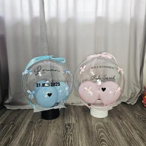 Op de afbeelding: Twee doorzichtige, ronde cadeau dozen met ballonnen erin. E&eacute;n doos heeft blauwe ballonnen, de naam "Louan" en de datum "31.M.2025". De andere doos heeft roze ballonnen en de tekst "WILL KOMMEN Baby Sarah". Beide hebben linten.
