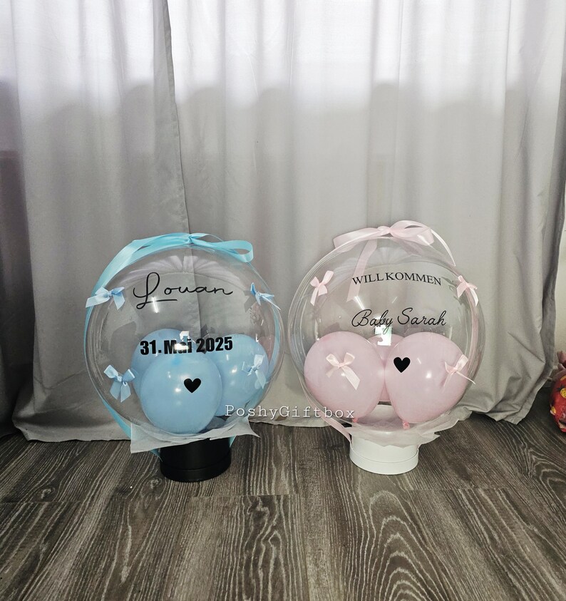Op de afbeelding: Twee doorzichtige, ronde cadeau dozen met ballonnen erin. E&eacute;n doos heeft blauwe ballonnen, de naam "Louan" en de datum "31.M.2025". De andere doos heeft roze ballonnen en de tekst "WILL KOMMEN Baby Sarah". Beide hebben linten.
