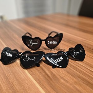 May include: Four black heart-shaped sunglasses with white text. One pair says "Sandra" and the other three say "Braut" and "TEAM".