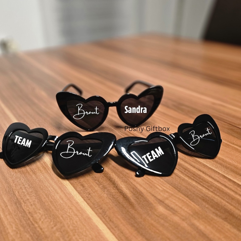 May include: Four black heart-shaped sunglasses with white text. One pair says "Sandra" and the other three say "Braut" and "TEAM".