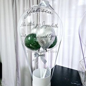 Op de afbeelding: Een doorzichtige ballon gevuld met kleinere groene en zilveren ballonnen, voorzien van de tekst 'Jalisia', 'Alles Liebe zur Jugendweihe' en 'Deine Oma und Opa' in zwart schrift. Het is bevestigd op een witte cilindrische voet met een zilveren lint.