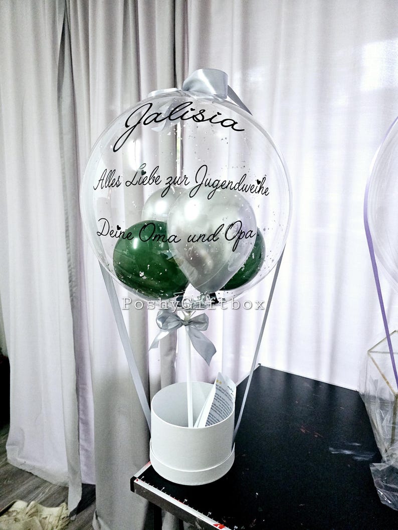 Op de afbeelding: Een doorzichtige ballon gevuld met kleinere groene en zilveren ballonnen, voorzien van de tekst 'Jalisia', 'Alles Liebe zur Jugendweihe' en 'Deine Oma und Opa' in zwart schrift. Het is bevestigd op een witte cilindrische voet met een zilveren lint.