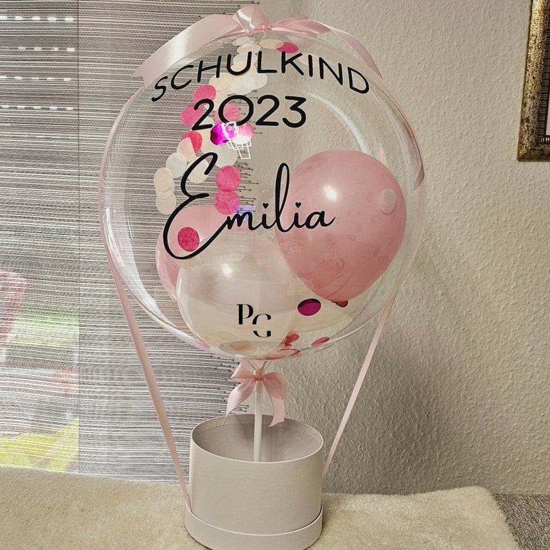 May include: A clear balloon shaped like a hot air balloon with pink, white, and confetti inside. The balloon has a black ribbon and a white box underneath. The balloon has the text "Schulkind 2023 Emilia" on it.