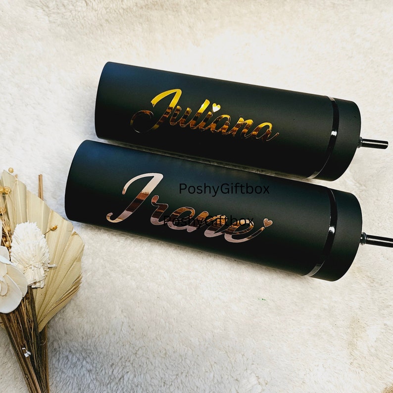 May include: Two black insulated tumblers with gold and rose gold vinyl lettering. The tumblers are personalized with the names "Juliana" and "Irene".