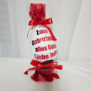 Peut inclure: Un &oelig;uf en chocolat blanc envelopp&eacute; de cellophane rouge avec un ruban rouge. L'&oelig;uf est d&eacute;cor&eacute; d'un texte noir qui dit "Zum Geburtstag alles Gute liebe Julia".