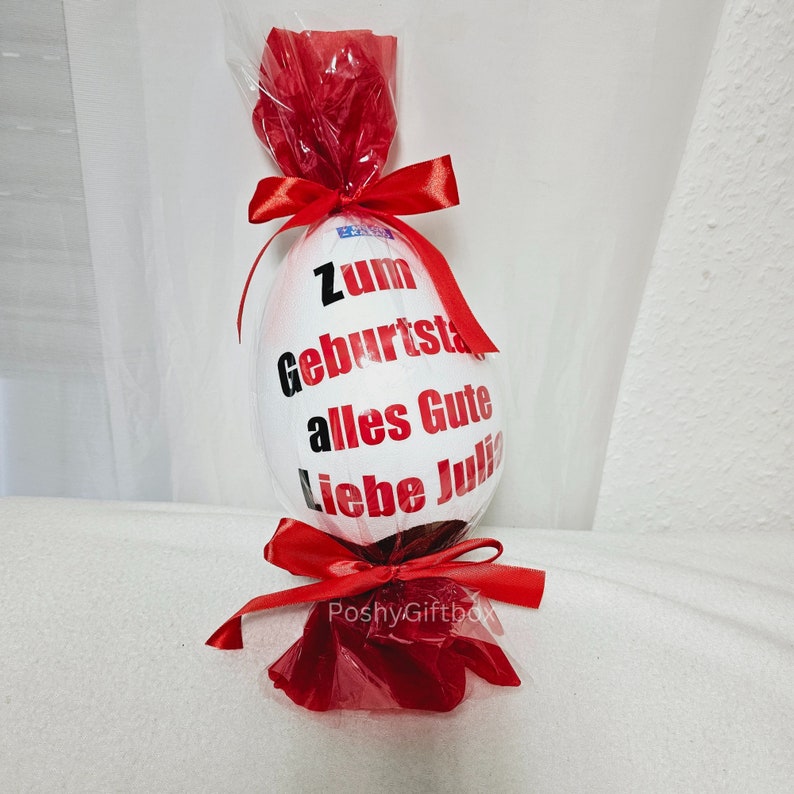 Peut inclure: Un &oelig;uf en chocolat blanc envelopp&eacute; de cellophane rouge avec un ruban rouge. L'&oelig;uf est d&eacute;cor&eacute; d'un texte noir qui dit "Zum Geburtstag alles Gute liebe Julia".