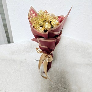 May include: A bouquet of chocolate truffles wrapped in gold foil, with dried baby's breath and a gold bow. The bouquet is wrapped in burgundy paper with gold accents and the text "PoshyGiftbox" is printed on the paper.