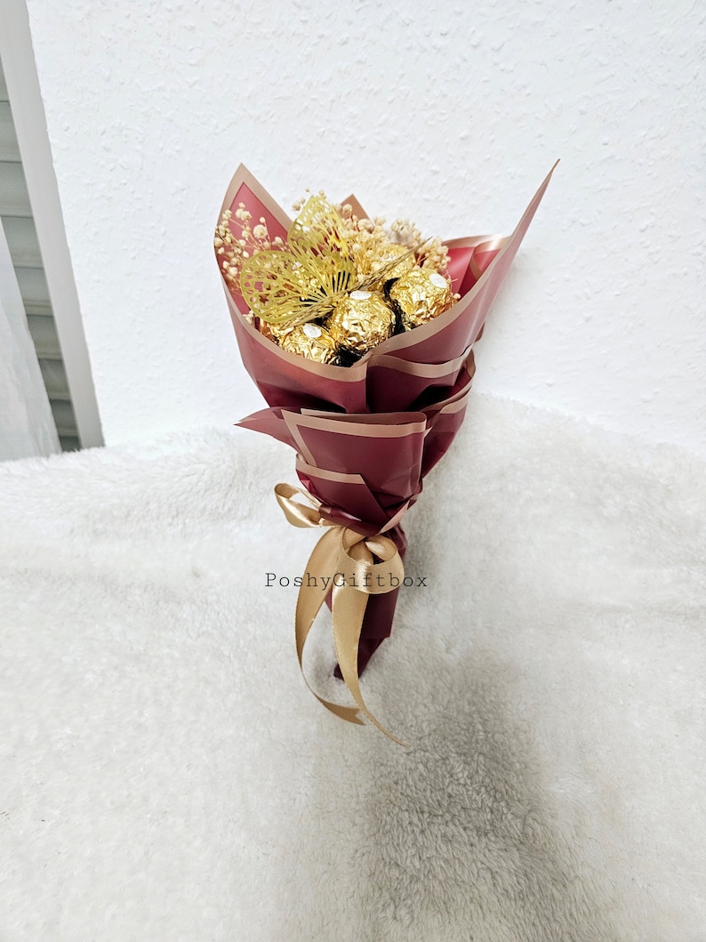 May include: A bouquet of chocolate truffles wrapped in gold foil, with dried baby's breath and a gold bow. The bouquet is wrapped in burgundy paper with gold accents and the text "PoshyGiftbox" is printed on the paper.