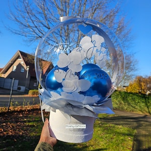 Op de afbeelding: Een heldere, bolvormige ballonnenopstelling met blauwe ballonnen binnenin, witte papieren uitsparingen en een witte basis met bloemmotief. De ballon is beschreven met namen en een datum. De basis heeft de tekst "Poshy Giftbox".