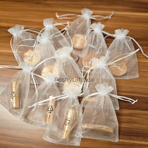 Pu&ograve; includere: Sacchetti di organza bianchi riempiti con piccoli cerchi di legno con nomi scritti su di essi. I sacchetti sono su una superficie di legno.