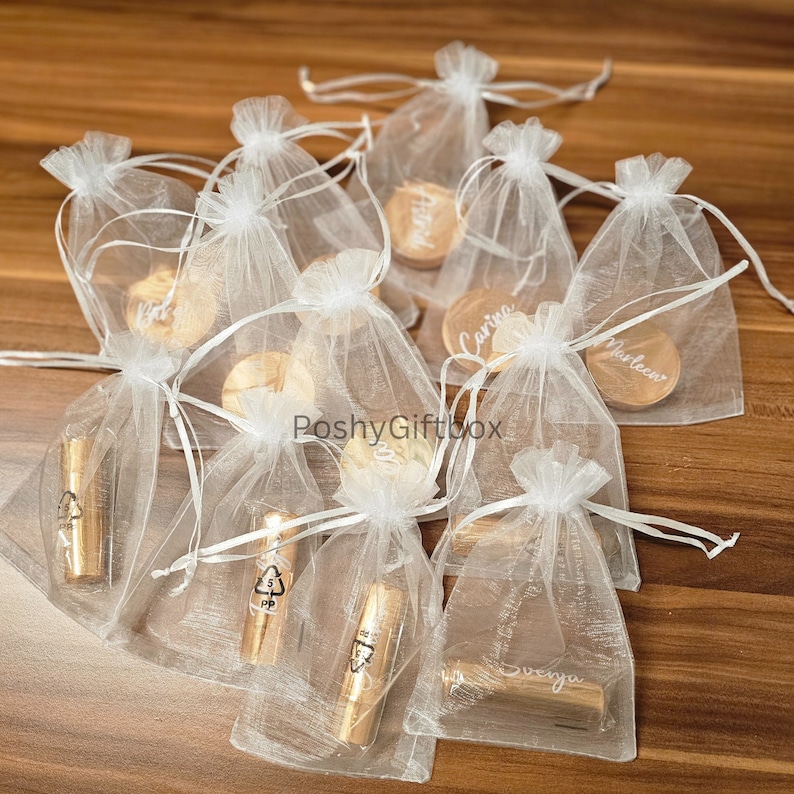 Pu&ograve; includere: Sacchetti di organza bianchi riempiti con piccoli cerchi di legno con nomi scritti su di essi. I sacchetti sono su una superficie di legno.