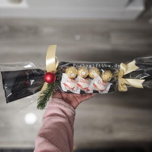 May include: A clear cellophane-wrapped gift cracker containing four gold-wrapped chocolates and three white-wrapped coconut candies. It is decorated with a red ornament, a small green pine sprig, and gold ribbons. The text Poshygiftbox.de is visible.