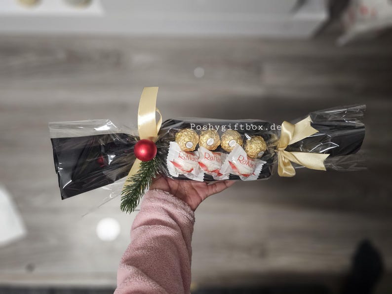 May include: A clear cellophane-wrapped gift cracker containing four gold-wrapped chocolates and three white-wrapped coconut candies. It is decorated with a red ornament, a small green pine sprig, and gold ribbons. The text Poshygiftbox.de is visible.
