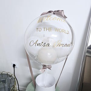 May include: A clear balloon with a white box underneath. The balloon has a gold ribbon and the words "Welcome to the World Anisa Imani" written on it.