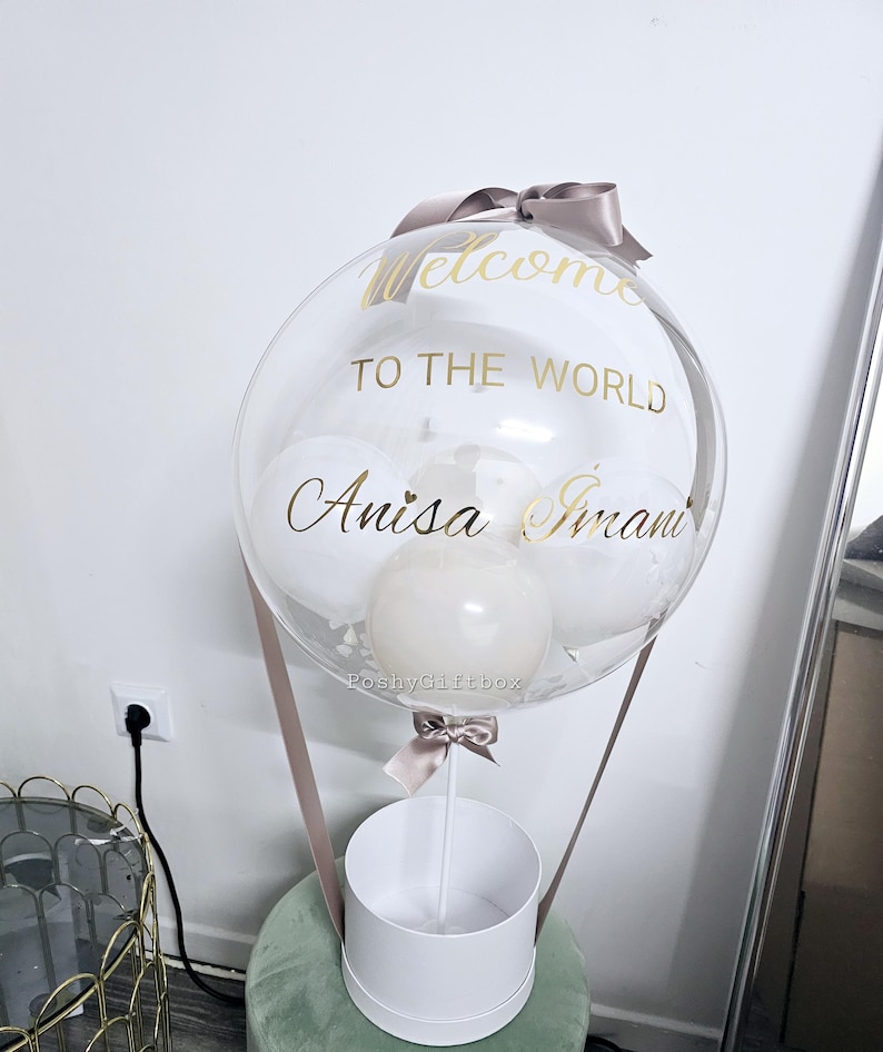 May include: A clear balloon with a white box underneath. The balloon has a gold ribbon and the words "Welcome to the World Anisa Imani" written on it.