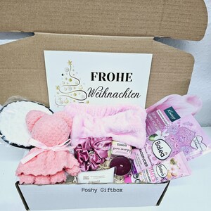 Peut inclure: Une bo&icirc;te cadeau rose remplie de divers articles, dont un ours en peluche rose, un bandeau rose, une bombe de bain rose, une bougie et une bouteille de lotion. La bo&icirc;te est d&eacute;cor&eacute;e d'un sapin de No&euml;l et des mots "Frohe Weihnachten" qui signifie "Joyeux No&euml;l" en allemand.