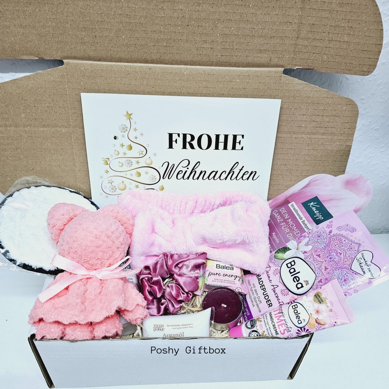 Peut inclure: Une bo&icirc;te cadeau rose remplie de divers articles, dont un ours en peluche rose, un bandeau rose, une bombe de bain rose, une bougie et une bouteille de lotion. La bo&icirc;te est d&eacute;cor&eacute;e d'un sapin de No&euml;l et des mots "Frohe Weihnachten" qui signifie "Joyeux No&euml;l" en allemand.