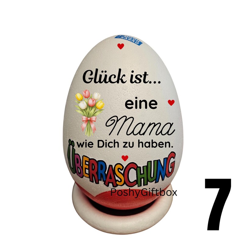 May include: A white egg-shaped decorative item with the text "Gl&uuml;ck ist... eine Mama wie Dich zu haben" and "&Uuml;BERRASCHUNG". It features a bouquet of flowers and red hearts. The item is on a white and red base.