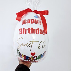 Może przedstawiać: Biały balon z napisem "Happy Birthday" w kolorze czerwonym i "Sweet 60" w kolorze złotym. Balon jest owinięty przezroczystym celofanem i zawiązany czerwoną wstążką. Widoczne jest r&oacute;wnież słowo "&Uuml;berraschung".
