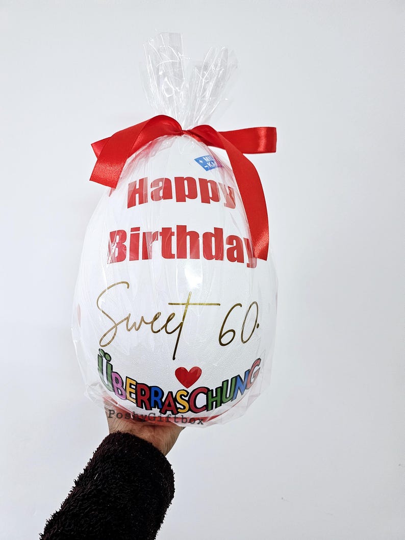 Może przedstawiać: Biały balon z napisem "Happy Birthday" w kolorze czerwonym i "Sweet 60" w kolorze złotym. Balon jest owinięty przezroczystym celofanem i zawiązany czerwoną wstążką. Widoczne jest r&oacute;wnież słowo "&Uuml;berraschung".