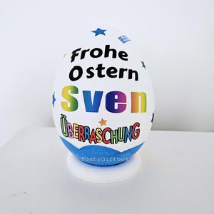 Op de afbeelding: Een wit paasei met een blauwe basis. Het ei is versierd met kleurrijke tekst die "Frohe Ostern Sven &Uuml;berraschung" en sterren zegt. De basis van het ei heeft de tekst "PoshyGiftbox" erop gedrukt.