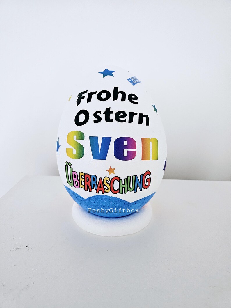 Op de afbeelding: Een wit paasei met een blauwe basis. Het ei is versierd met kleurrijke tekst die "Frohe Ostern Sven &Uuml;berraschung" en sterren zegt. De basis van het ei heeft de tekst "PoshyGiftbox" erop gedrukt.