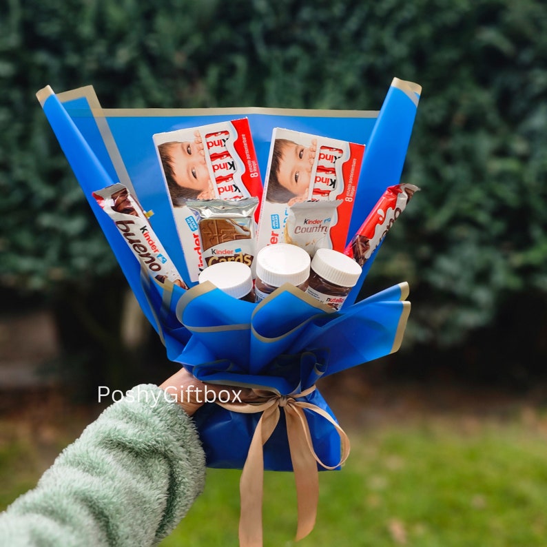 May include: A blue and gold gift bouquet with a variety of chocolate treats, including Kinder Bueno, Kinder Country, Kinder Cards, and Nutella. The bouquet is tied with a gold ribbon and has the text "PoshyGiftbox" printed on the bottom.