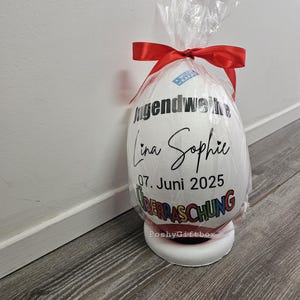 May include: A large, white egg-shaped gift wrapped in clear cellophane and tied with a red ribbon. The egg features black text that reads "Jugendweihe Lina Sophie 07. Juni 2025 &Uuml;BERRASCHUNG". The gift sits on a white base.