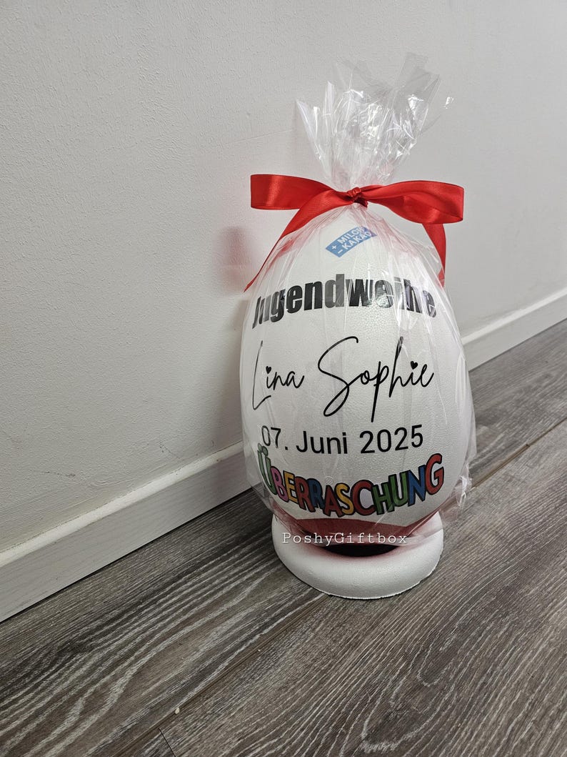 May include: A large, white egg-shaped gift wrapped in clear cellophane and tied with a red ribbon. The egg features black text that reads "Jugendweihe Lina Sophie 07. Juni 2025 &Uuml;BERRASCHUNG". The gift sits on a white base.