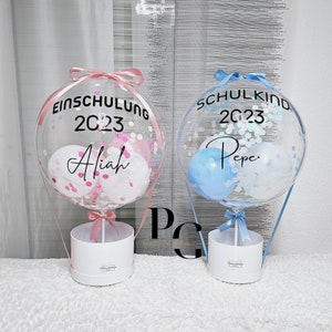 May include: Two clear balloon hot air balloons with confetti and small balloons inside. The balloons are tied to white boxes with pink and blue ribbons. The pink balloon has the text "Einschulung 2023 Alijah" and the blue balloon has the text "Schulkind 2023 Pepe."
