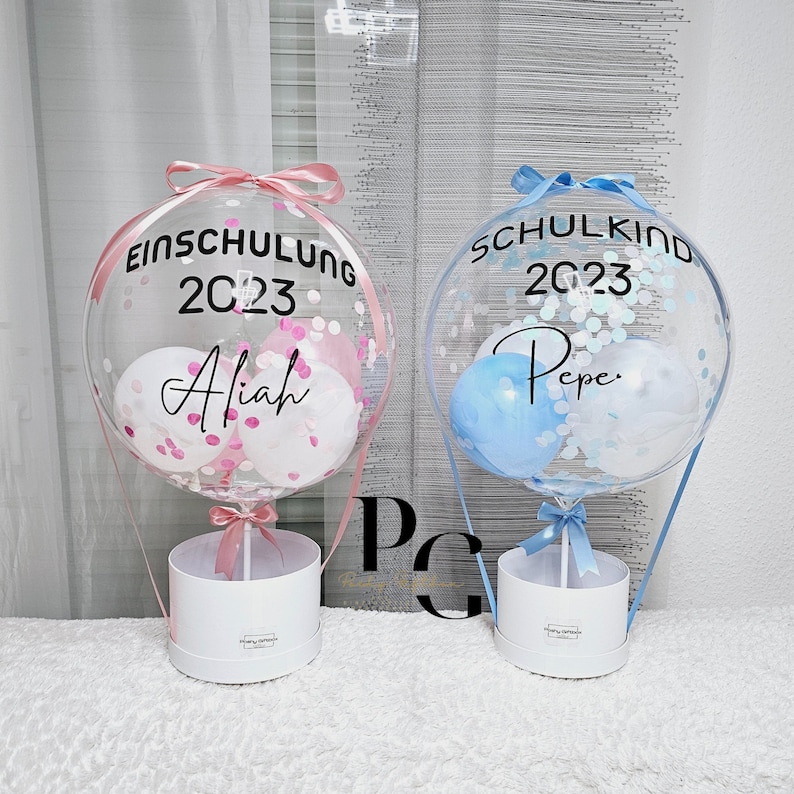 May include: Two clear balloon hot air balloons with confetti and small balloons inside. The balloons are tied to white boxes with pink and blue ribbons. The pink balloon has the text "Einschulung 2023 Alijah" and the blue balloon has the text "Schulkind 2023 Pepe."