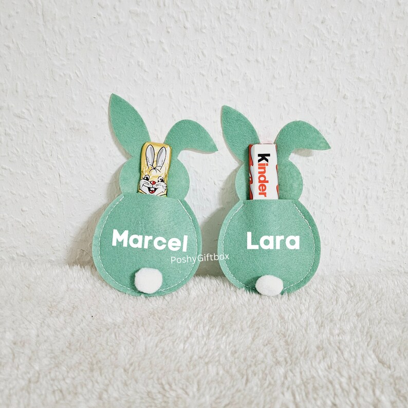 May include: Two green felt bunny-shaped Easter baskets with white pom-poms on the bottom. The baskets have pockets for treats and are personalized with the names "Marcel" and "Lara".