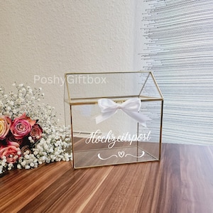 May include: A clear glass box with a gold frame and a white ribbon bow. The box has a slot on the top for inserting cards or envelopes. The box is labeled "Hochzeitspost" which means "wedding mail" in German.