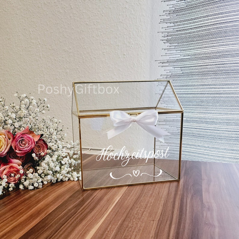 May include: A clear glass box with a gold frame and a white ribbon bow. The box has a slot on the top for inserting cards or envelopes. The box is labeled "Hochzeitspost" which means "wedding mail" in German.