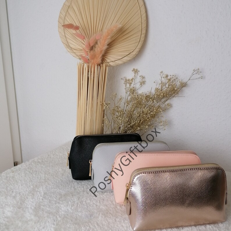 May include: Four cosmetic bags in different colors: black, gray, pink, and gold. The bags are made of a textured material and have a zipper closure. The bags are arranged in a row, with the black bag at the top and the gold bag at the bottom. The text "PoshyGiftbox" is visible in the image.