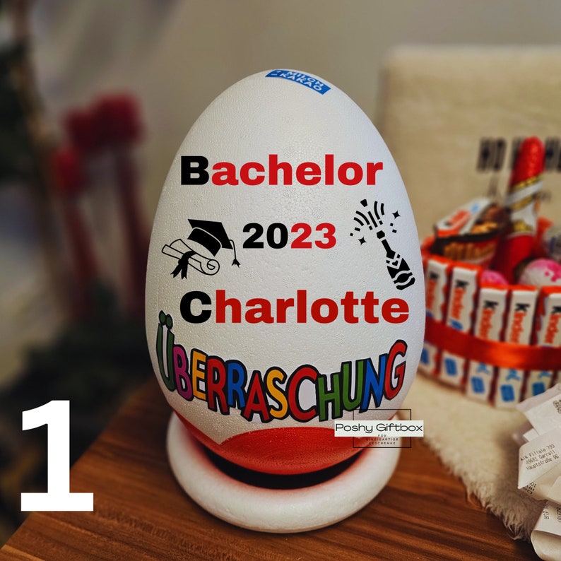 Może przedstawiać: Biały pojemnik w kształcie jajka z napisem "Bachelor 2023 Charlotte" i "Ueberraschung". Pojemnik jest ozdobiony czapką absolwenta, zwojem i petardą. Pojemnik stoi na białej podstawie.