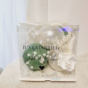 May include: A clear box with a white bow, containing white and green balloons, white flowers, and the text "JUST MARRIED Sabi & Keith". The box has a heart-shaped confetti design and a white rose.