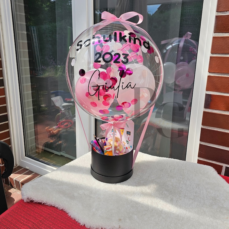 May include: A clear balloon shaped like a hot air balloon with pink and white confetti inside. The balloon is tied with a pink ribbon and has a black sign that says "Schulkind 2023 Ginia". The balloon is sitting on top of a black gift box filled with small gifts.
