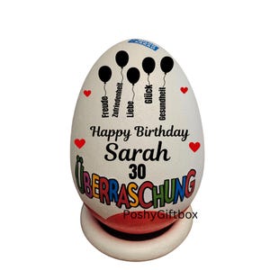 Może przedstawiać: Biały, dekoracyjny przedmiot w kształcie jajka z napisem "Happy Birthday Sarah 30" i niemieckim słowem "&Uuml;berraschung". Zawiera czarne grafiki balon&oacute;w i czerwone akcenty w kształcie serca. Przedmiot stoi na białej i czerwonej podstawie.