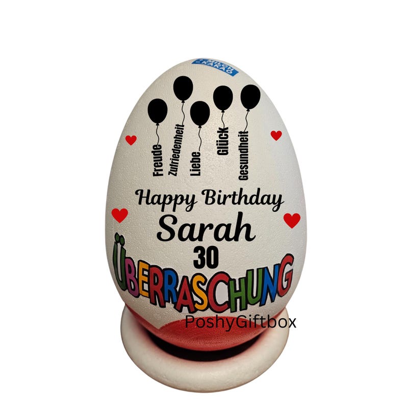 Może przedstawiać: Biały, dekoracyjny przedmiot w kształcie jajka z napisem "Happy Birthday Sarah 30" i niemieckim słowem "&Uuml;berraschung". Zawiera czarne grafiki balon&oacute;w i czerwone akcenty w kształcie serca. Przedmiot stoi na białej i czerwonej podstawie.