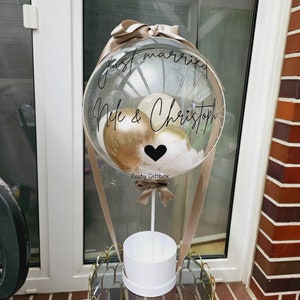 May include: A clear balloon with a white ribbon and a white box at the bottom. The balloon has gold and white balloons inside and the text "Just married Nele & Christoph" written on it. The balloon is tied to a stick with a brown ribbon.
