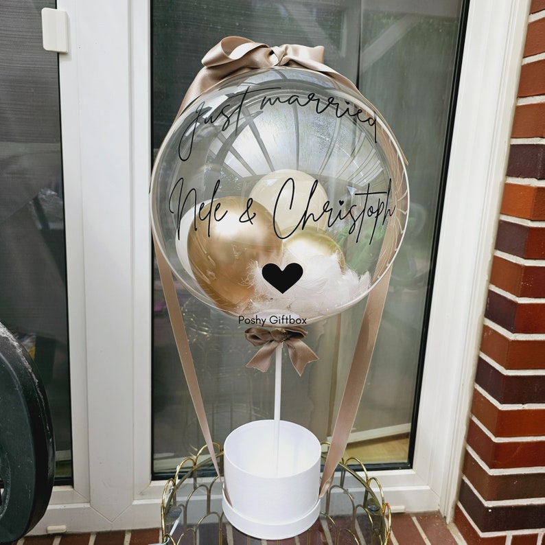 May include: A clear balloon with a white ribbon and a white box at the bottom. The balloon has gold and white balloons inside and the text "Just married Nele & Christoph" written on it. The balloon is tied to a stick with a brown ribbon.