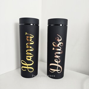 May include: Two black insulated tumblers with gold and rose gold lettering. The tumbler on the left says "Hanna" and the tumbler on the right says "Denise".