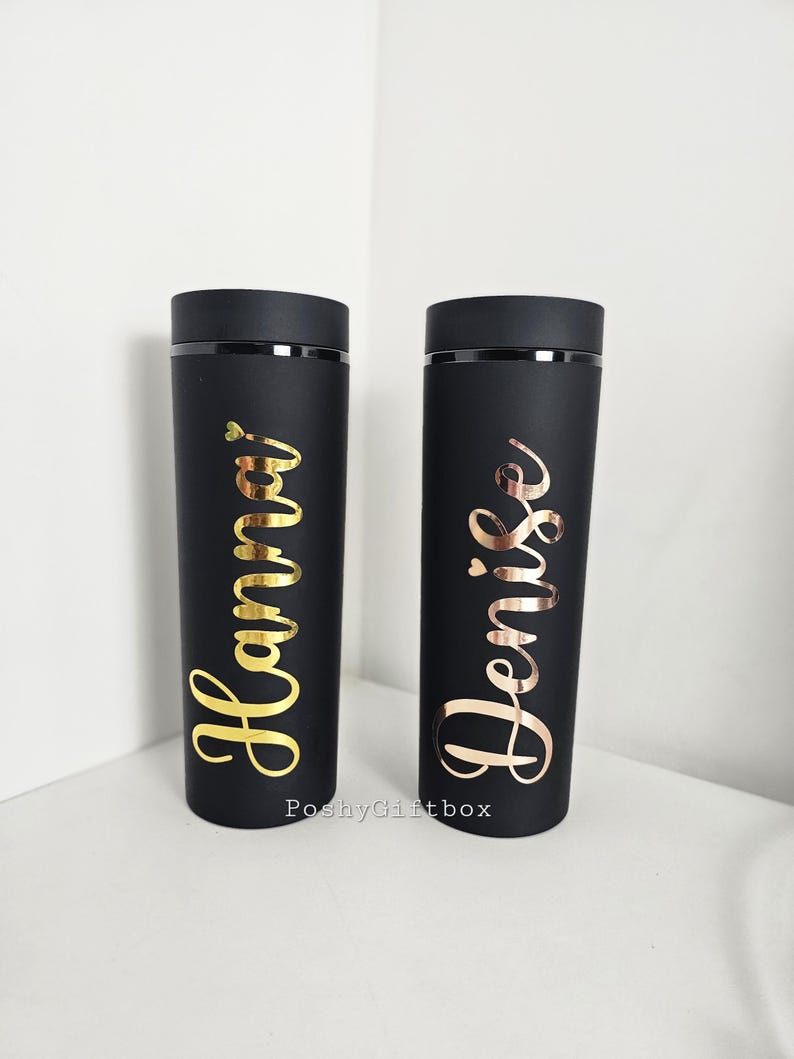 May include: Two black insulated tumblers with gold and rose gold lettering. The tumbler on the left says "Hanna" and the tumbler on the right says "Denise".