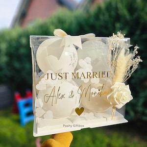 May include: A clear acrylic gift box with four white balloons, decorated with gold text that reads "JUST MARRIED" and the names "Alex & Monika". The box is adorned with a white ribbon, dried flowers, and a white rose.