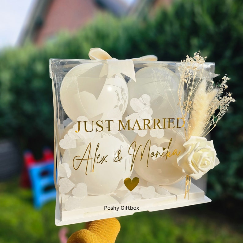 May include: A clear acrylic gift box with four white balloons, decorated with gold text that reads "JUST MARRIED" and the names "Alex & Monika". The box is adorned with a white ribbon, dried flowers, and a white rose.