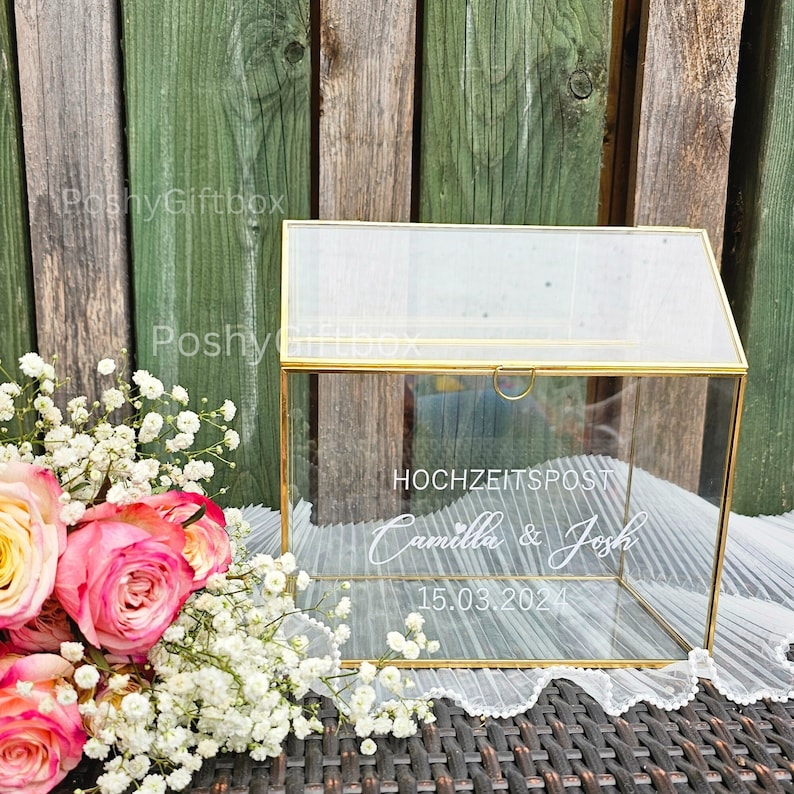 May include: A clear glass box with a gold frame and a hinged lid. The box is decorated with the text "HOCHZEITSPOST Camilla & Josh 15.03.2024" in white lettering.